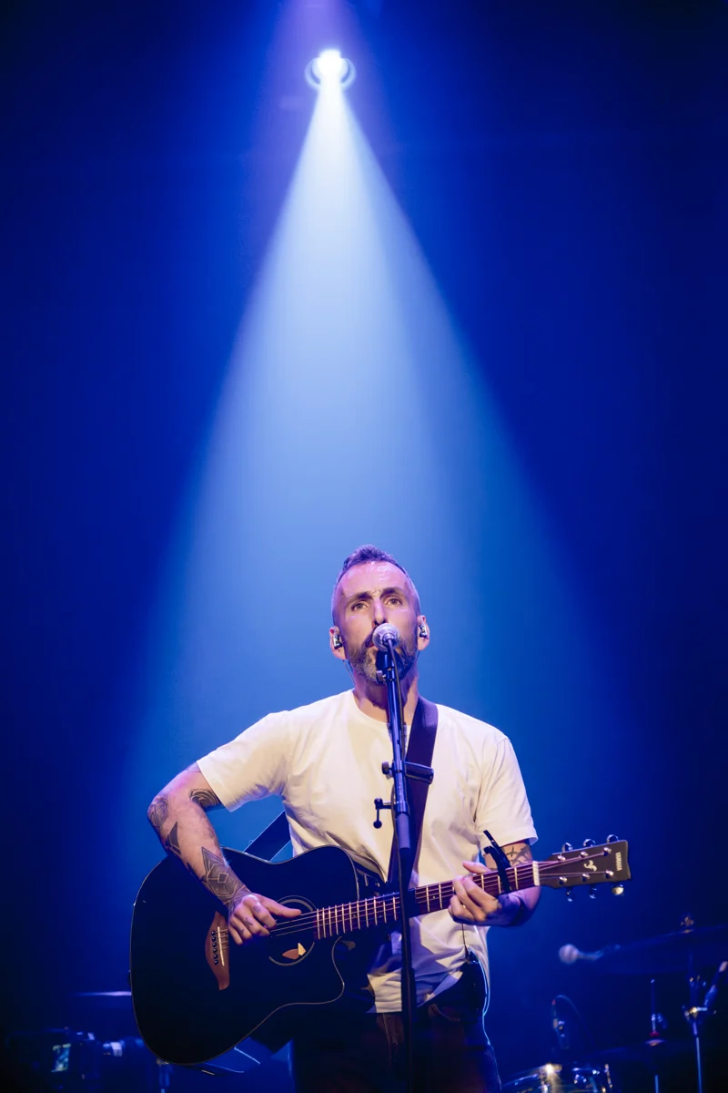 Nick Neo chante et joue de la guitare acoustique sous un faisceau bleu.